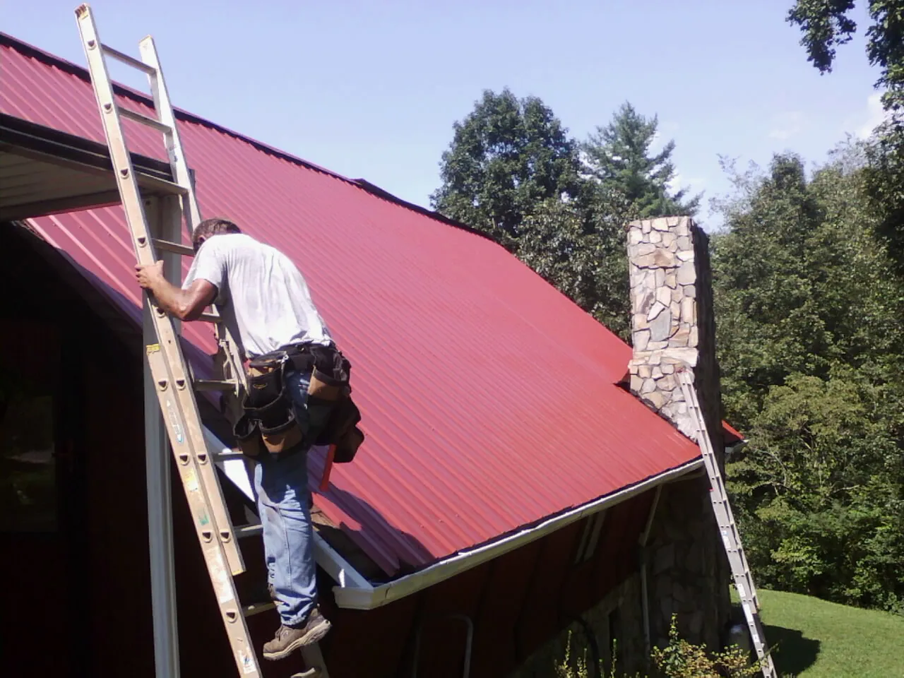 Roofer on a ladder beside a red metal roof and stone chimney — Soffit & Fascia Repair by Roofing Brothers in Gentryville
